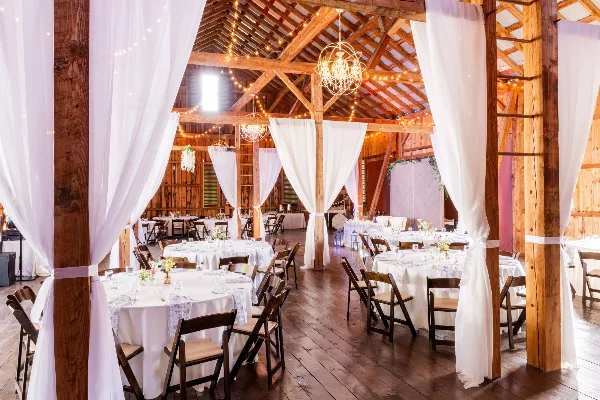 Interior view of a rustic wooden barn wedding venue featuring exposed timber beams, string lights, and round tables set for a reception.