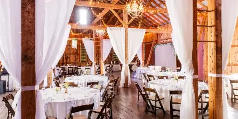 Interior view of a rustic wooden barn wedding venue featuring exposed timber beams, string lights, and round tables set for a reception.