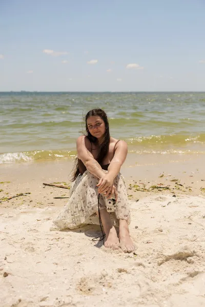 A woman sitting on a sandy beach holding a camera, with the ocean in the background.