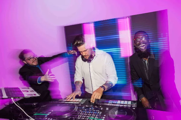 A DJ wearing headphones operates a Pioneer DJ controller and mixer while two colleagues stand behind him in a room illuminated by purple and blue stage lighting.