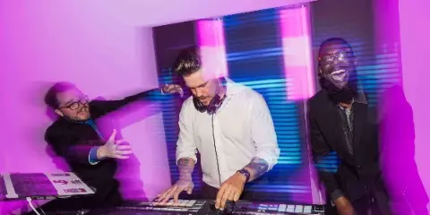 A DJ wearing headphones operates a Pioneer DJ controller and mixer while two colleagues stand behind him in a room illuminated by purple and blue stage lighting.