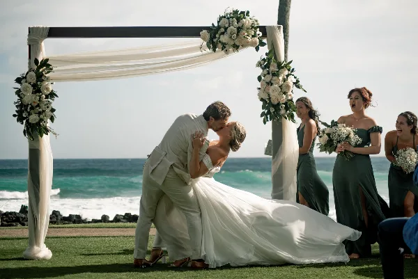 A bride and groom share a kiss under a floral arch during a beach wedding ceremony with bridesmaids in the background.