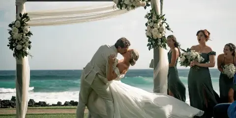A bride and groom share a kiss under a floral arch during a beach wedding ceremony with bridesmaids in the background.