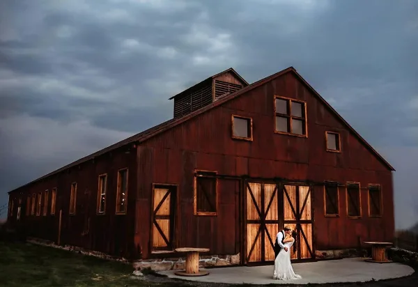 A red wooden barn with a couple standing in front of the main doors under a cloudy sky.