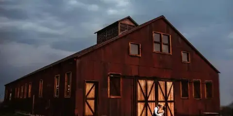A red wooden barn with a couple standing in front of the main doors under a cloudy sky.