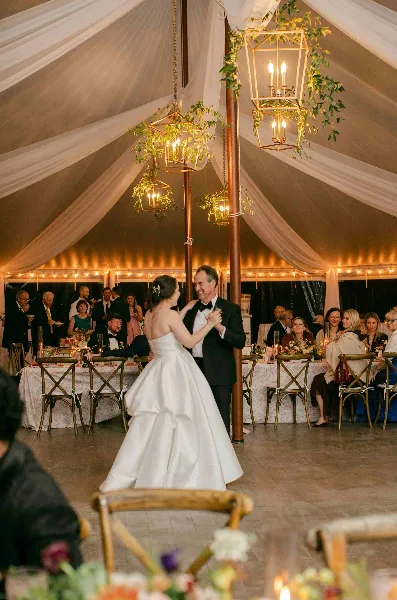A bride and groom dancing at a wedding reception inside a tent decorated with white fabric draping, hanging lanterns, and floral arrangements.