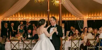 A bride and groom dancing at a wedding reception inside a tent decorated with white fabric draping, hanging lanterns, and floral arrangements.