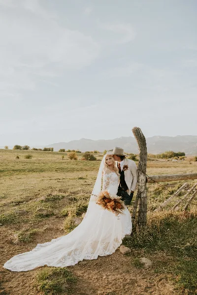 A bride and groom posing together next to a wooden fence in a grassy field with mountains in the distance.