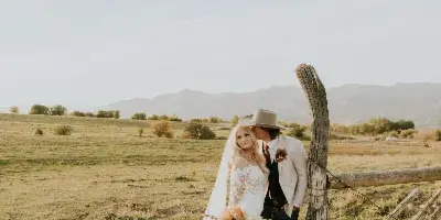 A bride and groom posing together next to a wooden fence in a grassy field with mountains in the distance.