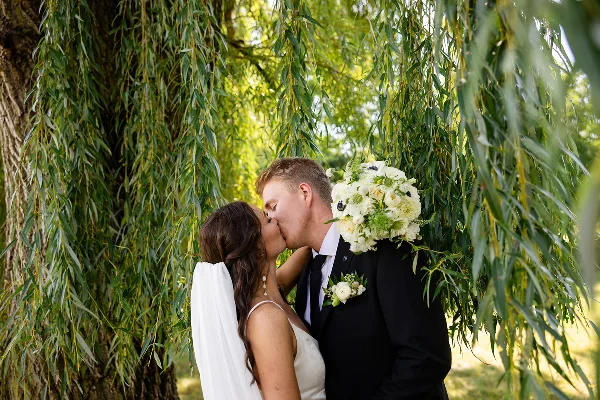 A bride and groom kissing under a weeping willow tree, with the bride holding a white bouquet.