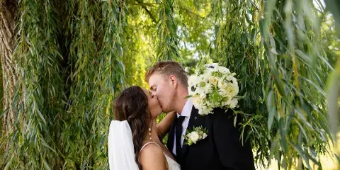 A bride and groom kissing under a weeping willow tree, with the bride holding a white bouquet.