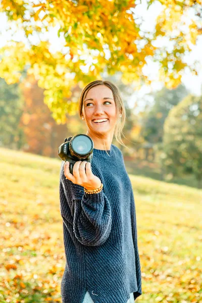 A smiling woman holding a professional camera outdoors in an autumn setting.