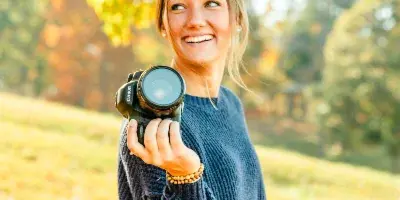 A smiling woman holding a professional camera outdoors in an autumn setting.