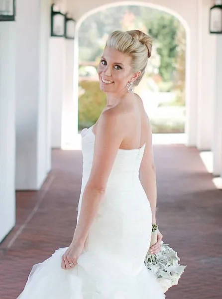 Bride in a white strapless gown with an elegant updo and polished makeup, holding a bouquet in a white arched corridor.
