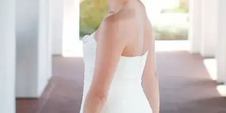 Bride in a white strapless gown with an elegant updo and polished makeup, holding a bouquet in a white arched corridor.