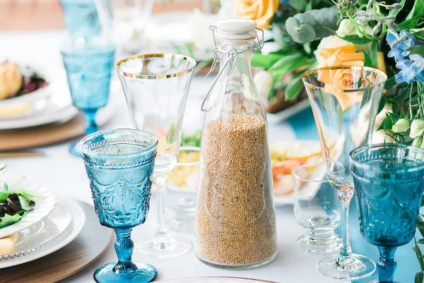 A close-up of a wedding table setting featuring blue glassware, a glass bottle of grain, and a floral arrangement with yellow roses and blue flowers.