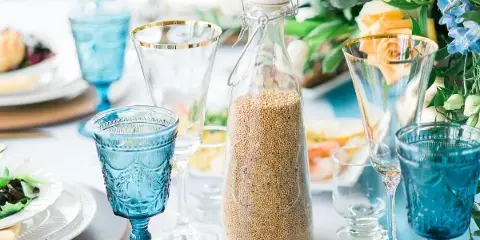 A close-up of a wedding table setting featuring blue glassware, a glass bottle of grain, and a floral arrangement with yellow roses and blue flowers.