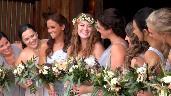A bride wearing a floral crown and white dress smiling with her bridesmaids, who are holding bouquets of white and peach flowers.