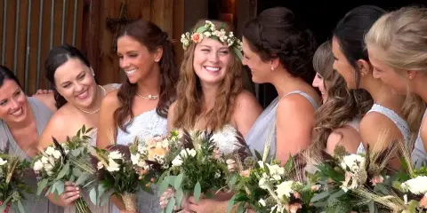 A bride wearing a floral crown and white dress smiling with her bridesmaids, who are holding bouquets of white and peach flowers.