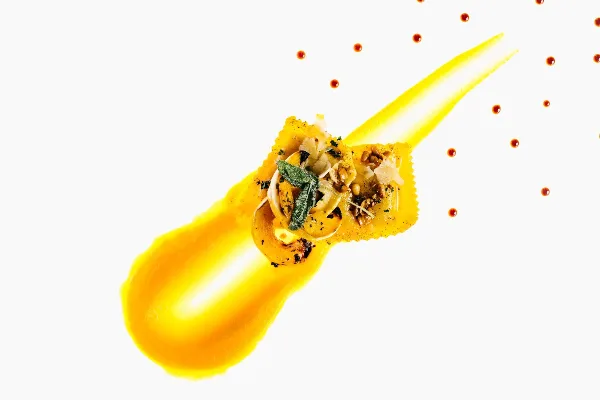 A professionally plated ravioli dish with yellow sauce and garnish on a white background.