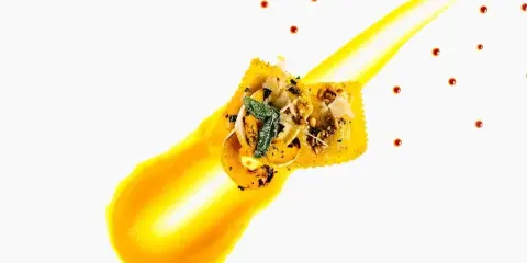 A professionally plated ravioli dish with yellow sauce and garnish on a white background.