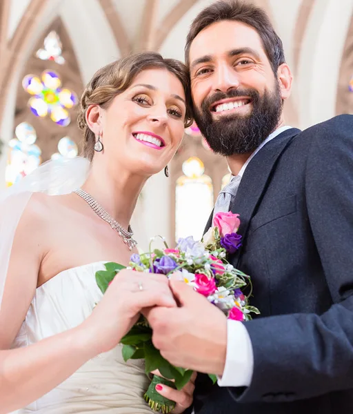 A smiling bride and groom posing together in a church setting, with the bride holding a colorful bouquet.