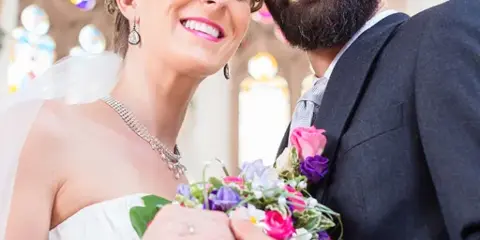 A smiling bride and groom posing together in a church setting, with the bride holding a colorful bouquet.