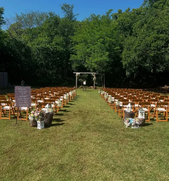 Outdoor wedding ceremony setup with rows of wooden chairs and a floral arch in a wooded clearing.