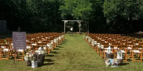Outdoor wedding ceremony setup with rows of wooden chairs and a floral arch in a wooded clearing.