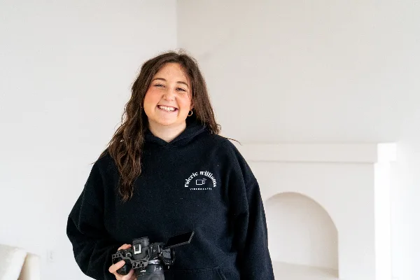 A smiling woman holding a professional video camera, wearing a black hoodie with the Valerie Williams Videography logo.