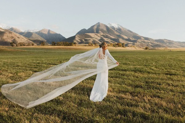 A bride in a white wedding dress and long veil standing in a grassy field with mountains in the background.