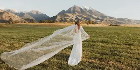 A bride in a white wedding dress and long veil standing in a grassy field with mountains in the background.