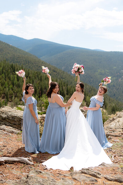 A bride and four bridesmaids in blue dresses holding bouquets, posing on a rocky mountain overlook.