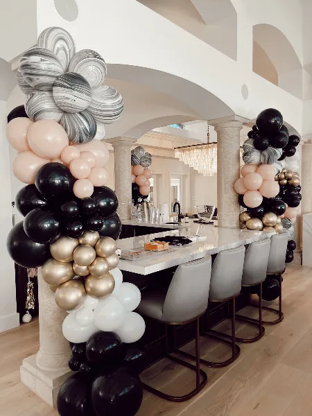 A luxury balloon installation featuring black, gold, white, and marbled balloons arranged around a kitchen island and columns.