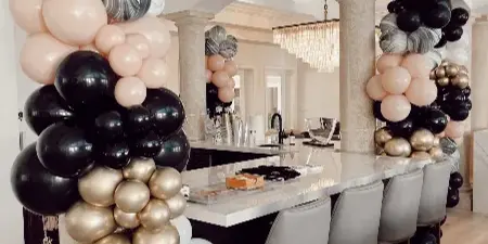 A luxury balloon installation featuring black, gold, white, and marbled balloons arranged around a kitchen island and columns.