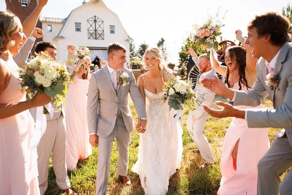 A bride and groom holding hands and smiling as they walk through a cheering wedding party in a grassy field.