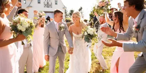 A bride and groom holding hands and smiling as they walk through a cheering wedding party in a grassy field.