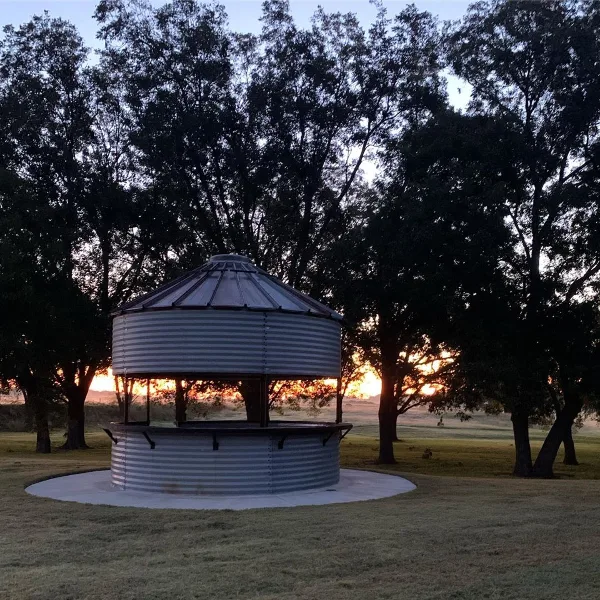 A round metal gazebo with a corrugated roof and bench seating, located on a grassy lawn surrounded by trees at dusk.