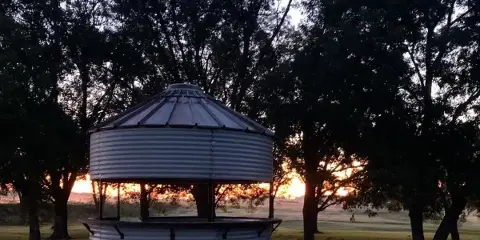 A round metal gazebo with a corrugated roof and bench seating, located on a grassy lawn surrounded by trees at dusk.