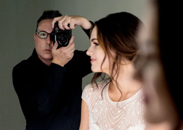 A photographer holding a camera takes a portrait of a woman in a white dress.
