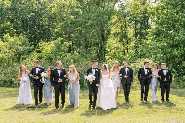 A wedding party consisting of a bride, groom, and attendants posing outdoors on a lawn with trees in the background.