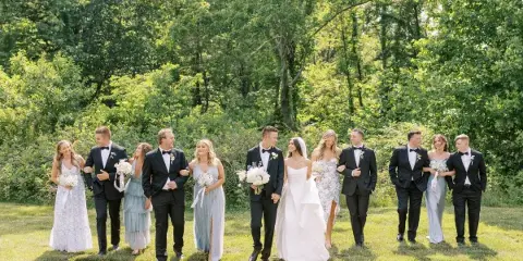 A wedding party consisting of a bride, groom, and attendants posing outdoors on a lawn with trees in the background.