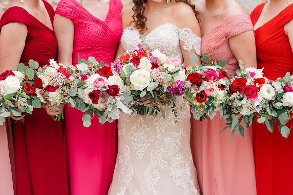 A bride and bridesmaids in red and pink dresses holding colorful floral bouquets with roses and eucalyptus.