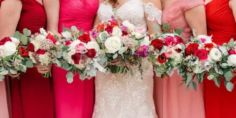A bride and bridesmaids in red and pink dresses holding colorful floral bouquets with roses and eucalyptus.