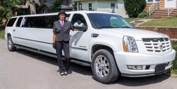 A professional chauffeur in a suit and hat standing next to a white stretch limousine.