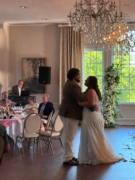A bride and groom share a first dance in a ballroom while a DJ operates a laptop and audio equipment in the background.