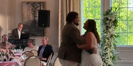 A bride and groom share a first dance in a ballroom while a DJ operates a laptop and audio equipment in the background.