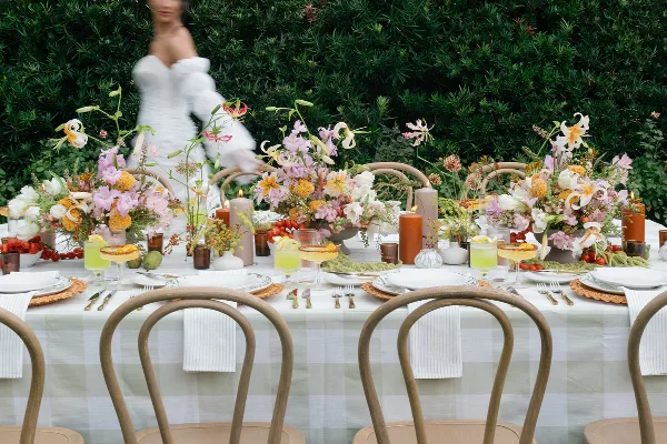 A wedding reception table decorated with lush floral centerpieces, candles, and place settings against a green hedge backdrop.
