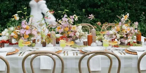 A wedding reception table decorated with lush floral centerpieces, candles, and place settings against a green hedge backdrop.