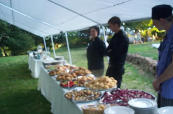 Outdoor catering event with a long table of food and staff under a white tent.